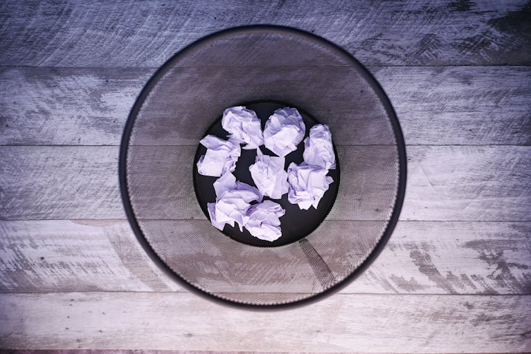 A top-down view of crumpled papers inside a black trash can on wooden flooring.