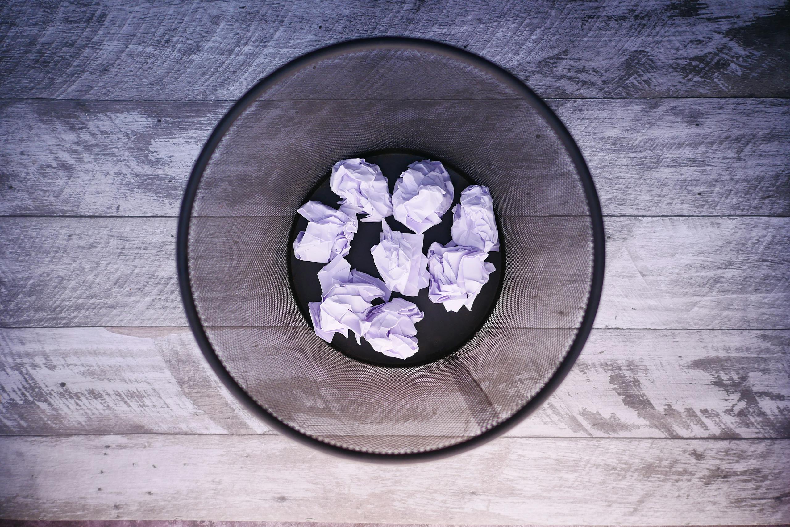 A top-down view of crumpled papers inside a black trash can on wooden flooring.